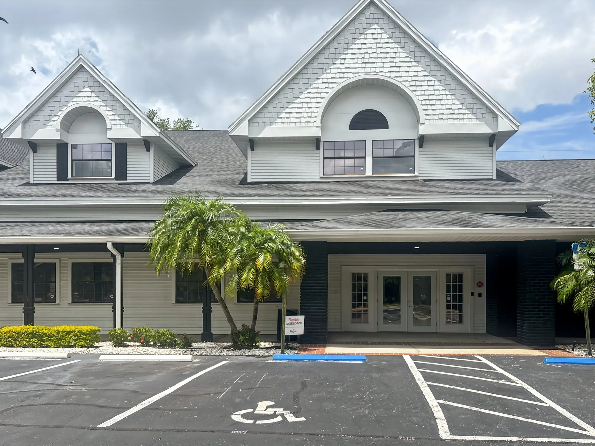 The white, gabled West building at 300 E Bay Dr with palm trees and parking out front