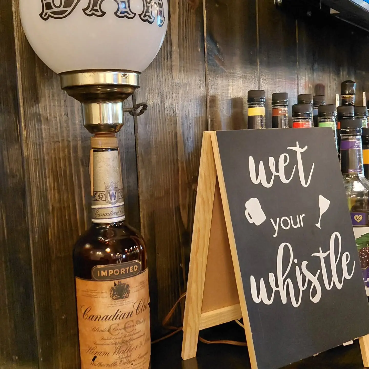 A “wet your whistle” chalkboard sign beside a vintage bottle lamp at the bar